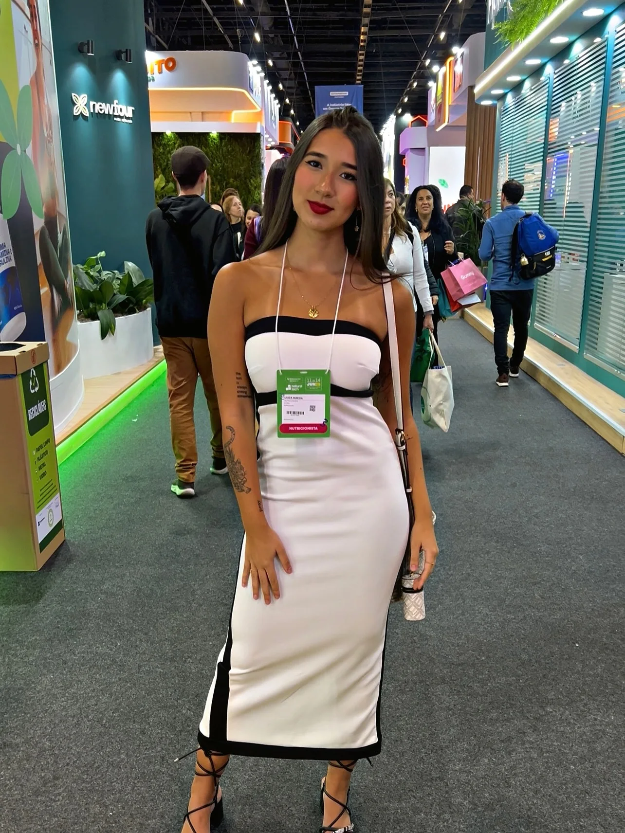 Luiza Maeda com crachá em um corredor de feira do setor de nutrição, de vestido branco e preto