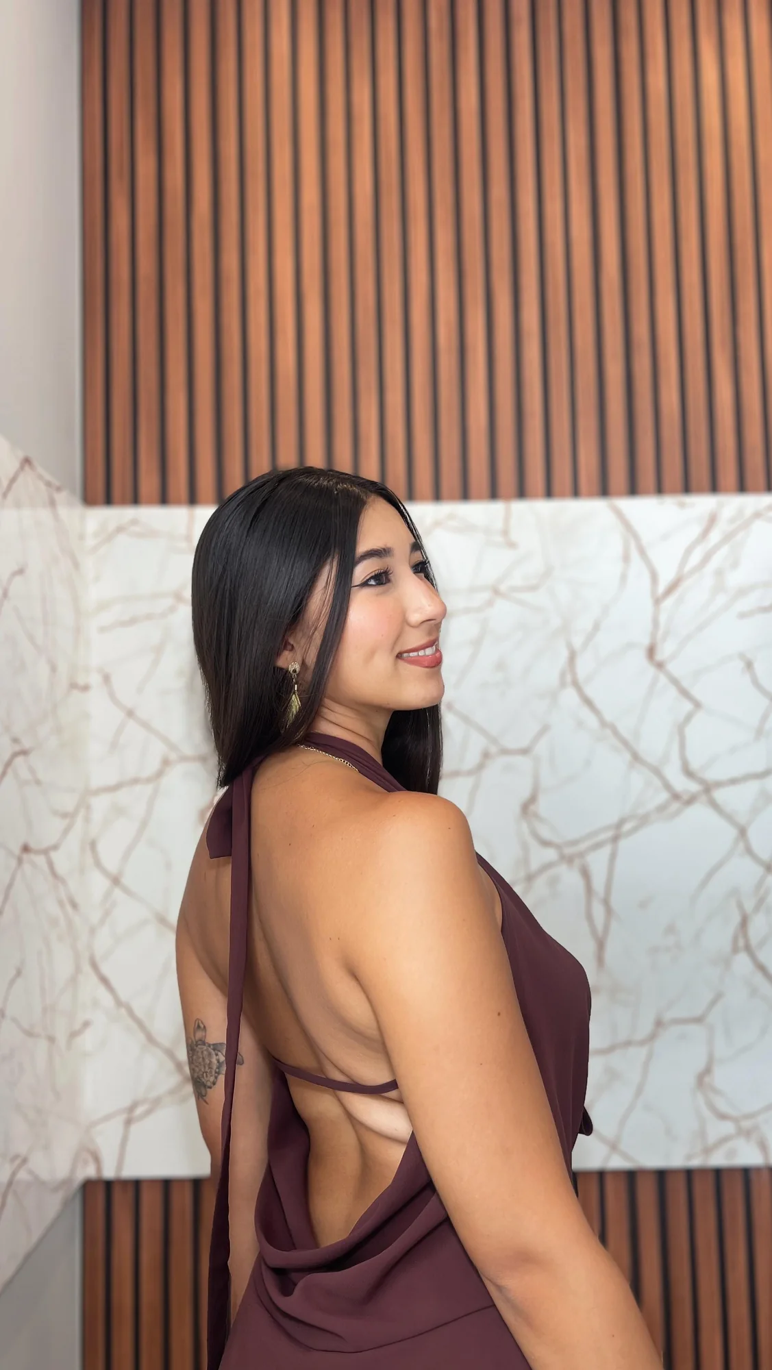 Luiza Maeda de perfil, olhando por cima do ombro, com vestido bordô de costas abertas e parede de madeira ao fundo