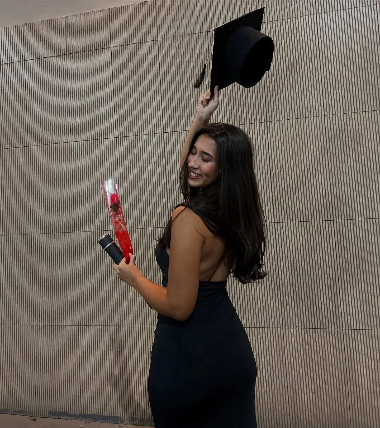 Luiza Maeda de costas, com vestido preto, jogando o capelo de formatura para cima