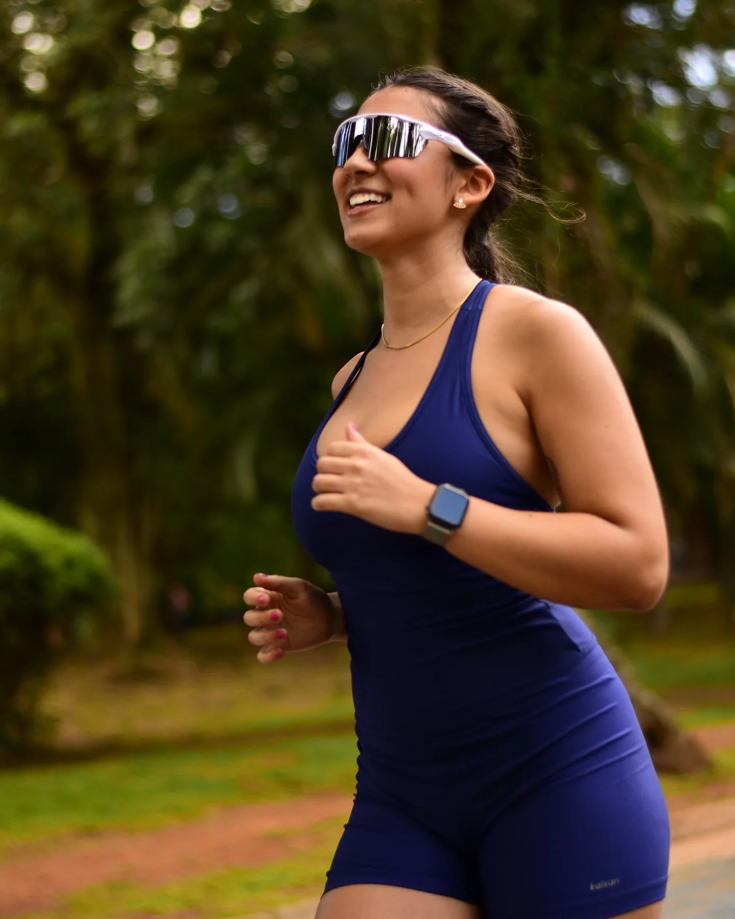 Luiza Maeda correndo em um parque, de macacão azul-marinho, óculos esportivos e sorriso aberto