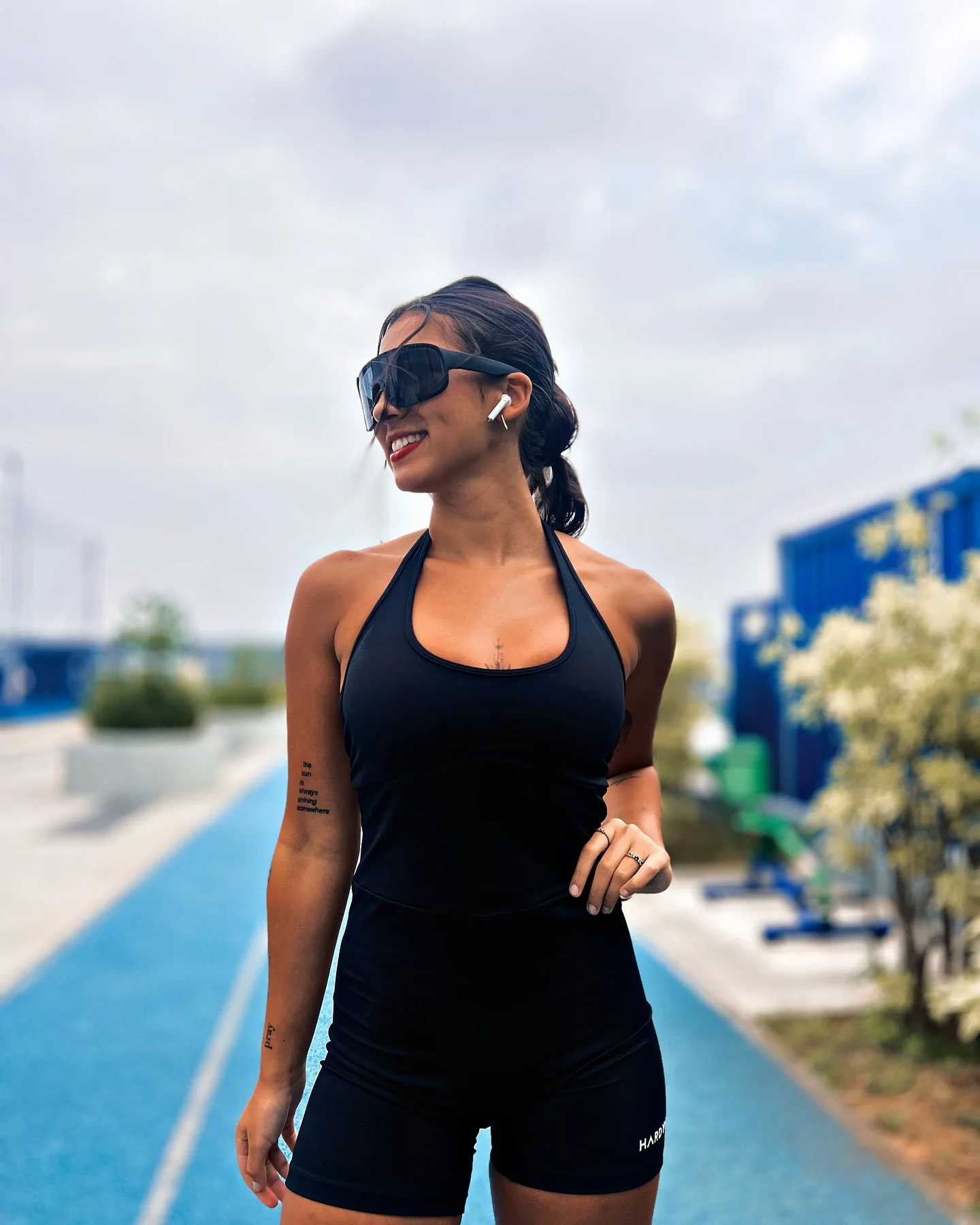 Luiza Maeda de óculos escuros e macacão preto ao lado de uma pista de corrida azul, sorrindo