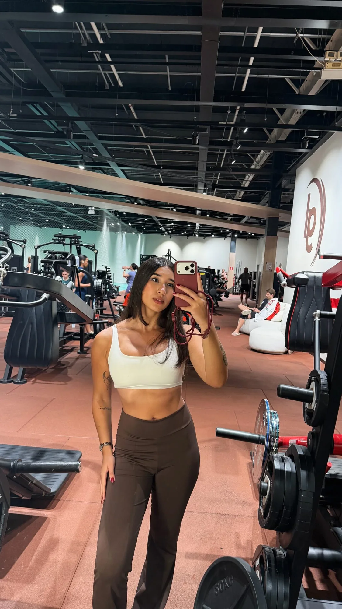 Luiza Maeda de top branco e calça marrom em pé no salão de uma academia