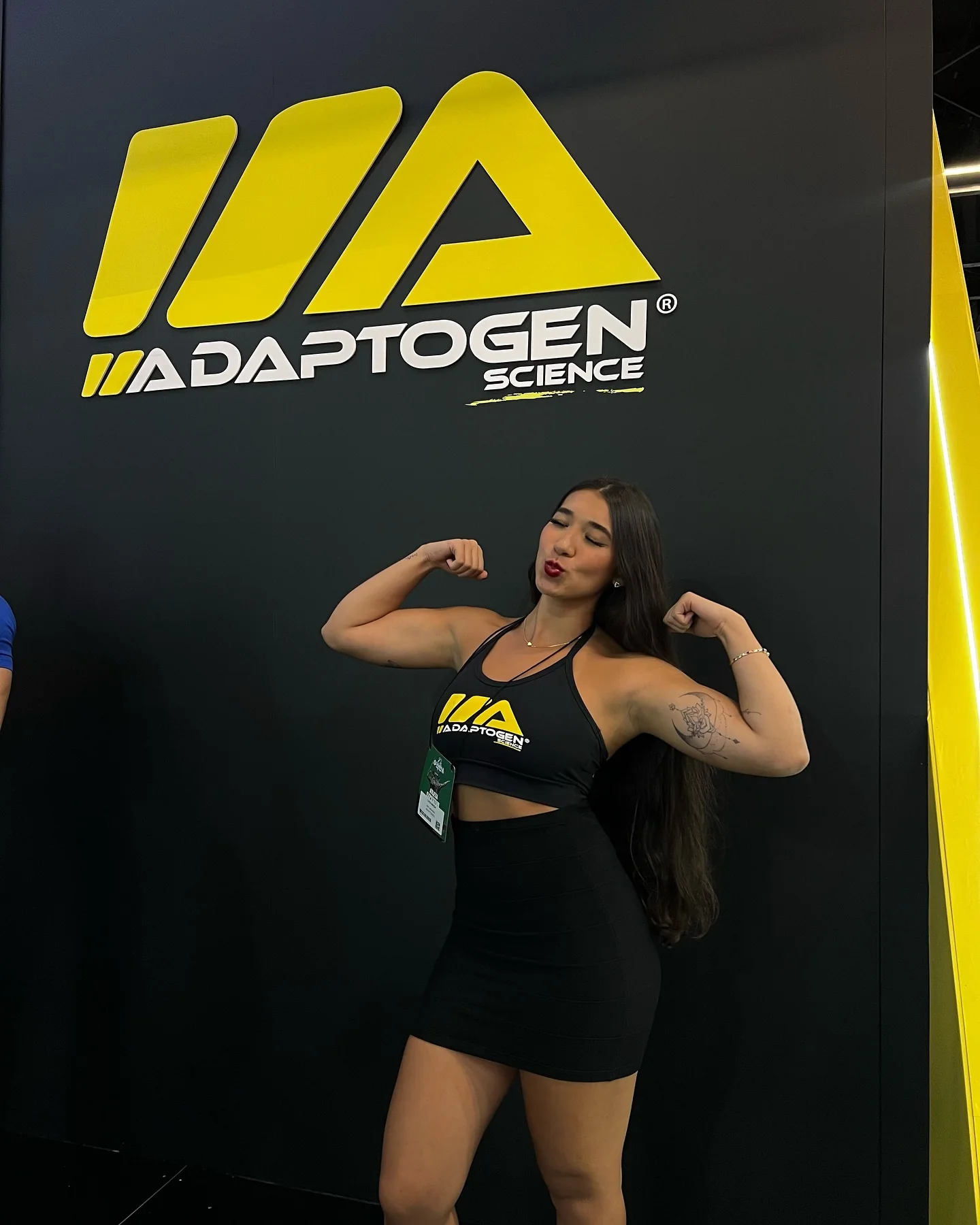 Luiza Maeda flexionando os braços diante do painel amarelo e preto da Adaptogen Science