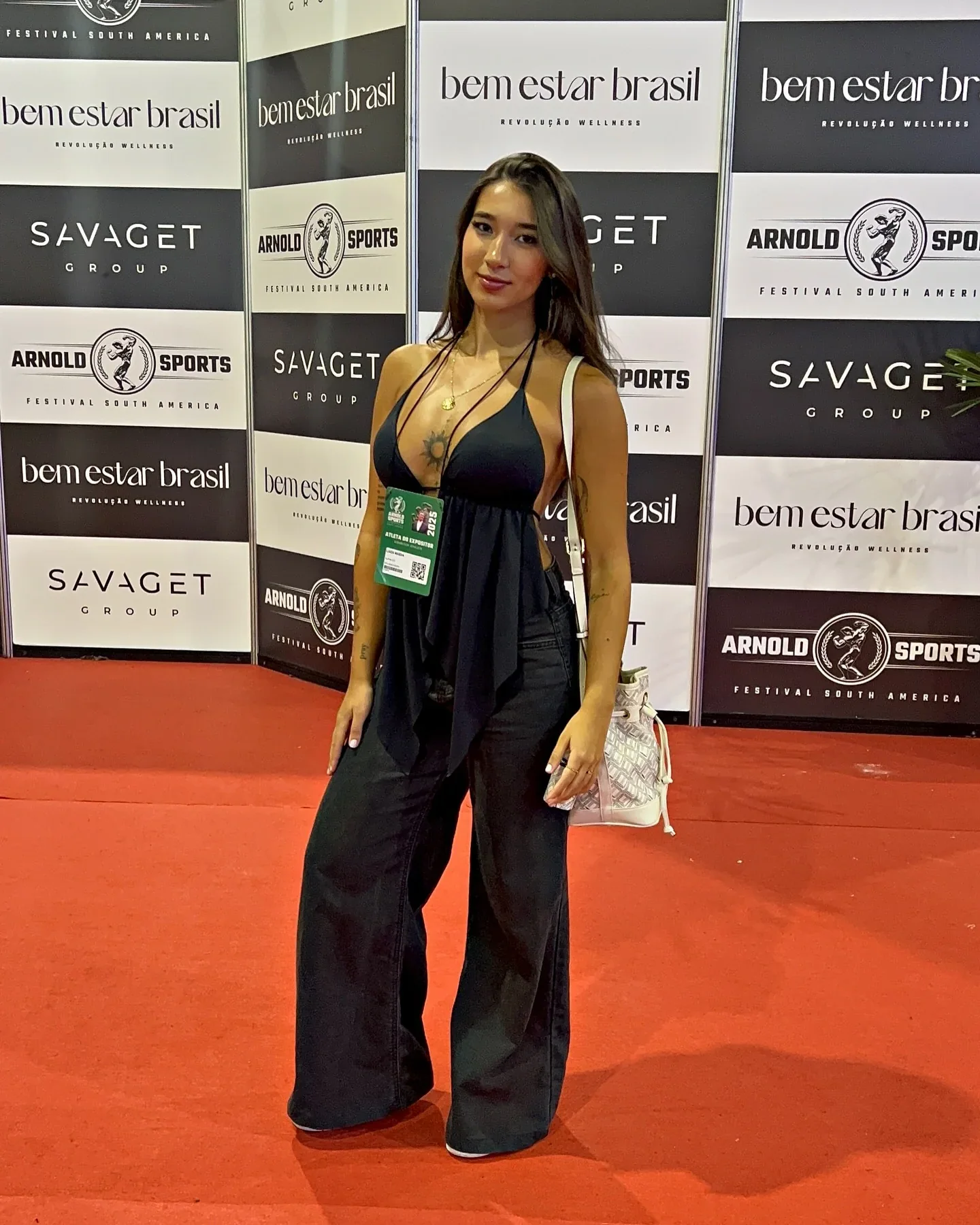 Luiza Maeda de macacão preto diante de um painel de patrocinadores em evento fitness
